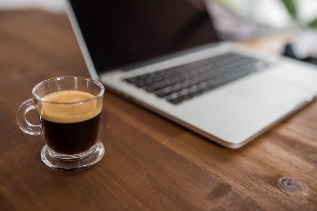 A glass cup of espresso sitting beside an open MacBook on a wooden desk