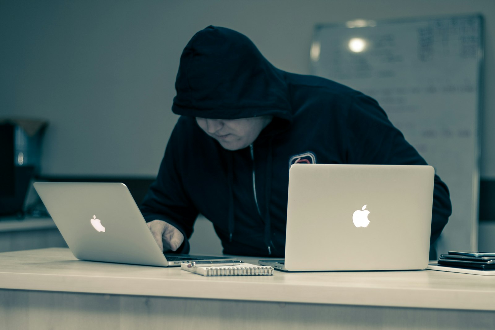 A hooded figure working between two Apple laptops, illustrating online scams